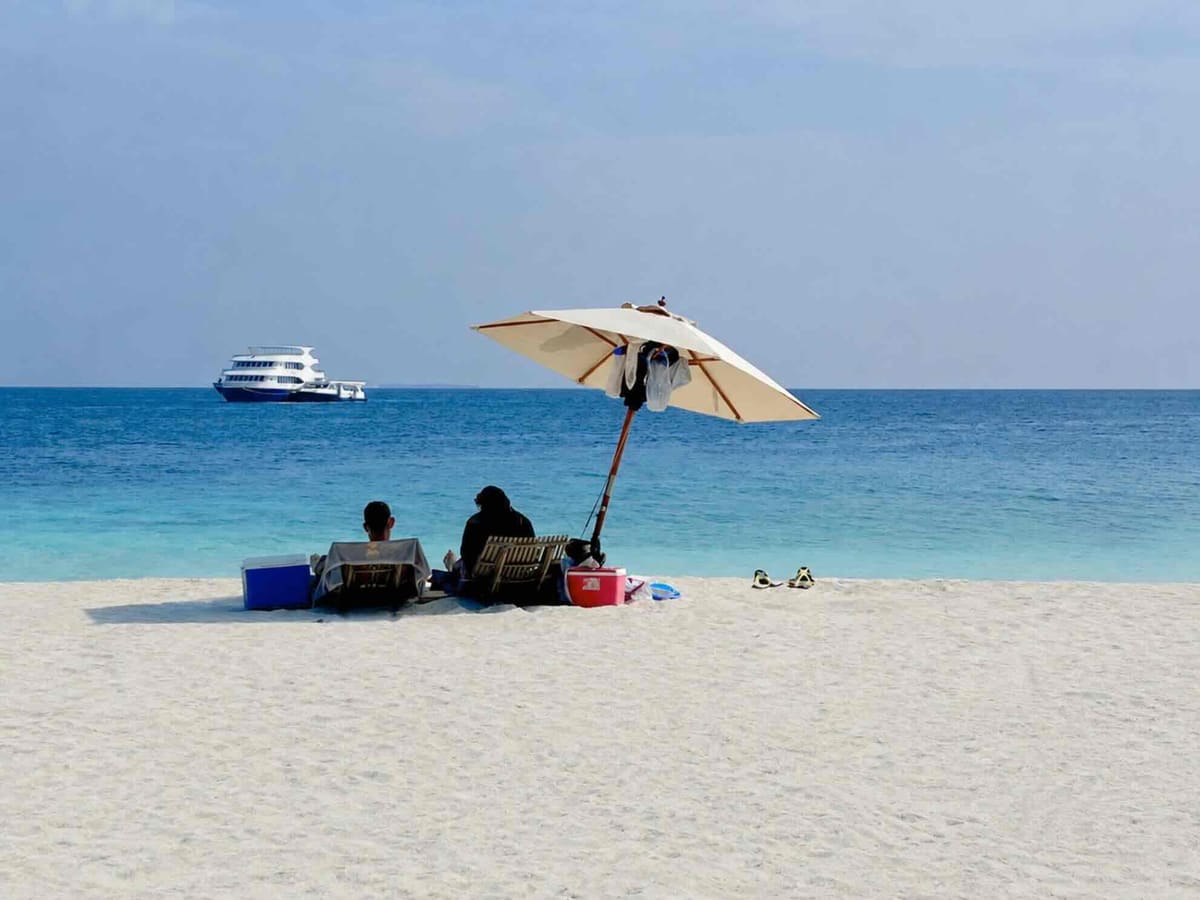 Ein Paar sitzt unter einem Sonnenschirm an einem einsamen Strand, das Safarischiff liegt vor Anker im Hintergrund